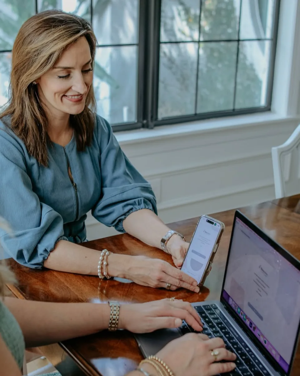 An agency owner checking her booking calendar on a phone and a laptop at her kitchen table