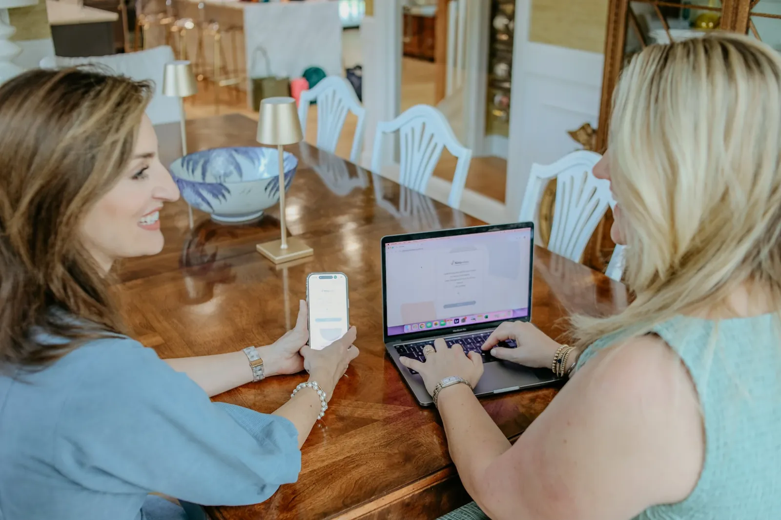 Two agency owners smiling at a table while reviewing their Sittysites booking app on a phone and a laptop