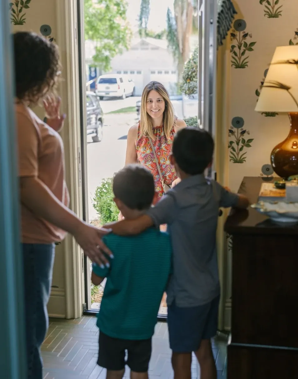 A mother and her two sons welcoming a smiling sitter at their front door